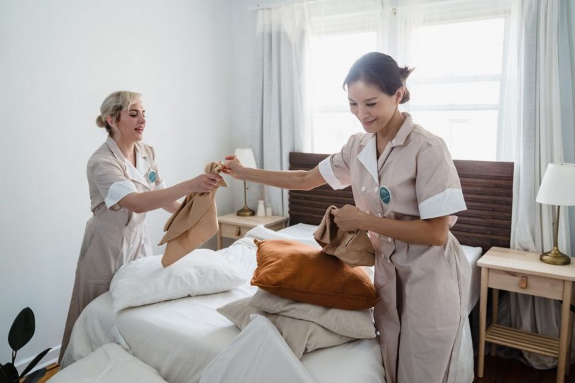 Técnico uniformizado aplicando controle de pragas em quarto de hotel com cama e cortina ao fundo
